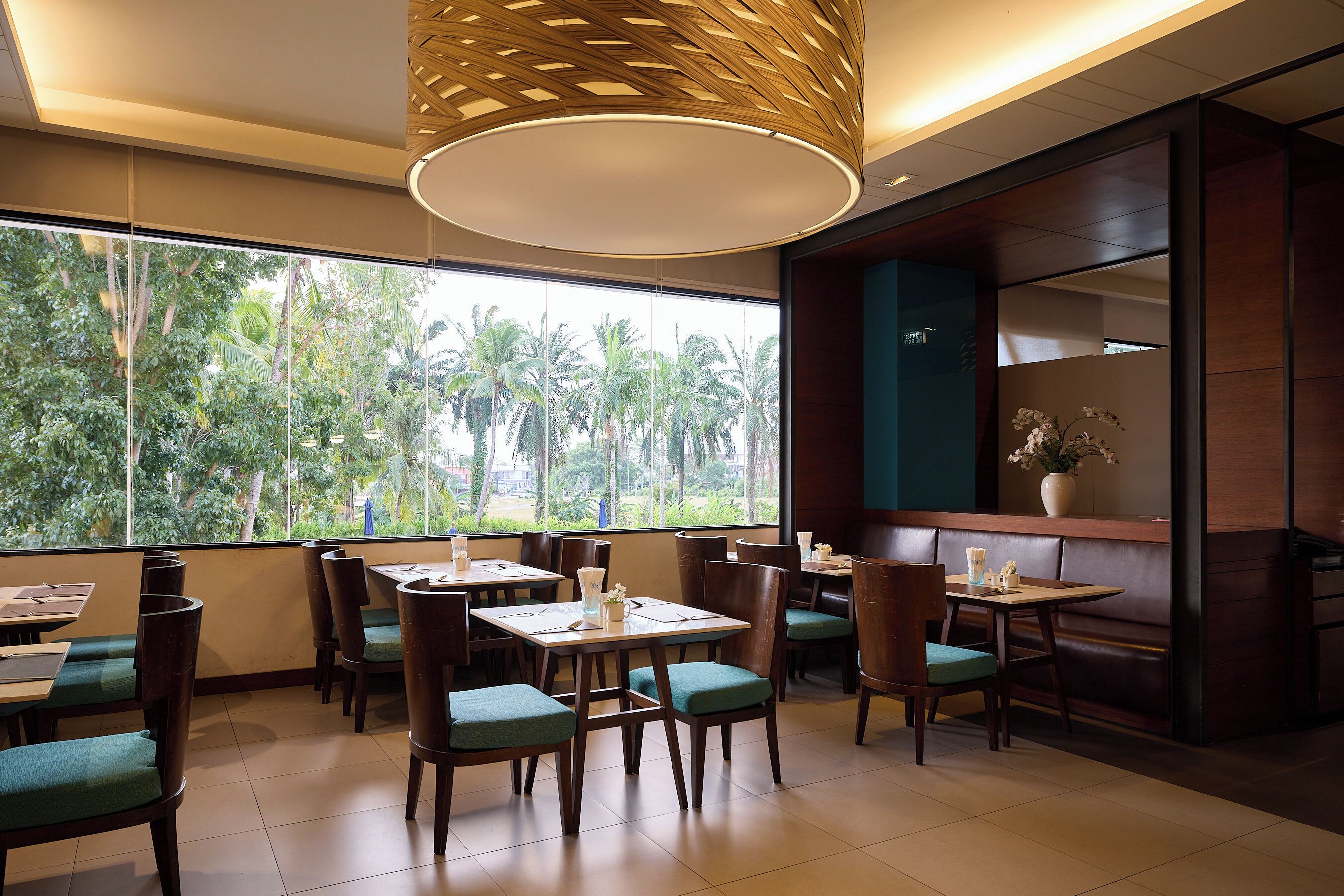 A restaurant with wooden tables and chairs, large windows with a view of palm trees, and a modern ceiling design.