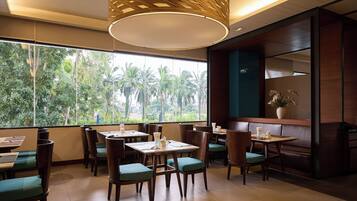 A restaurant with wooden tables and chairs, large windows with a view of palm trees, and a modern ceiling design.