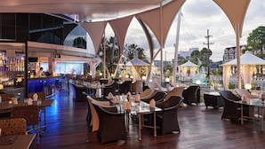 An outdoor restaurant with a bar, wicker furniture, and a view of the cityscape.