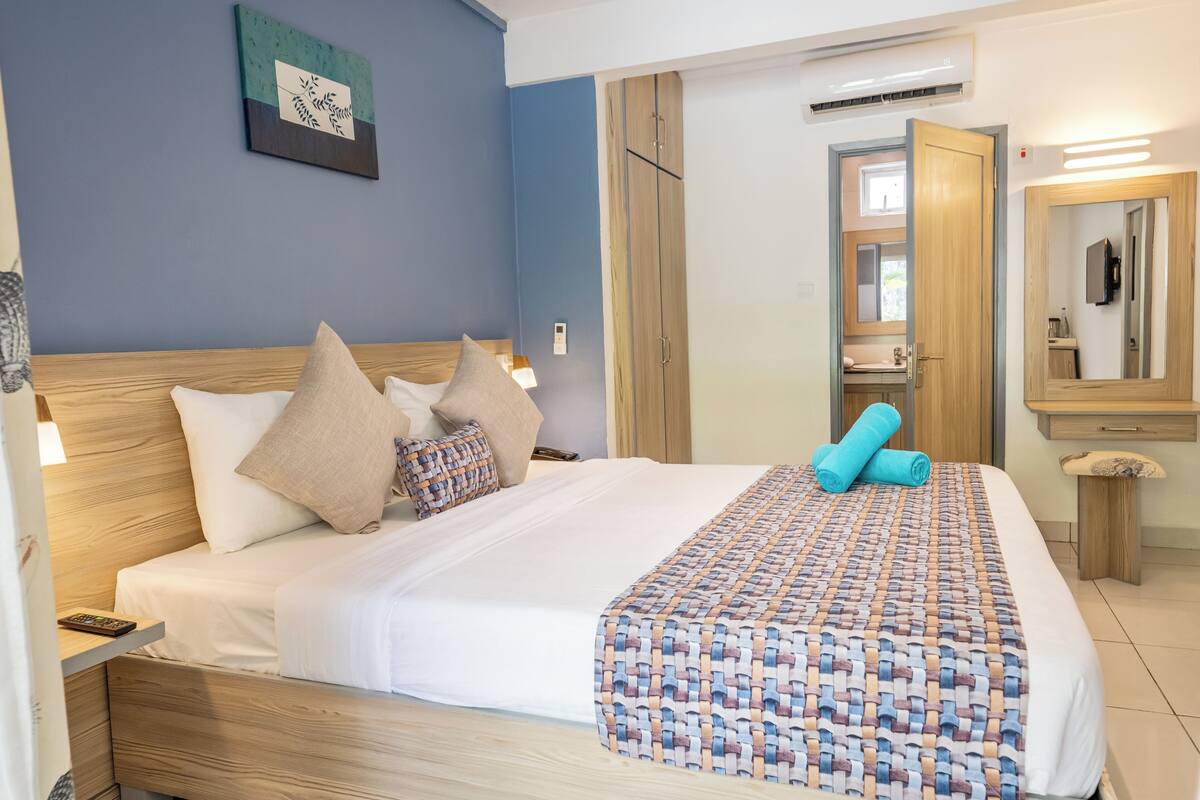 A neatly made bed with a patterned blanket, a wooden headboard, and several pillows. There is a wooden wardrobe with a mirror and a small table beside the bed.