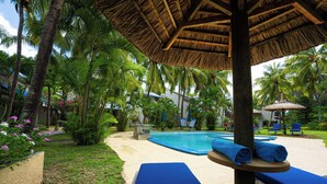 Un espace piscine avec des chaises longues, un parasol en chaume et des palmiers.