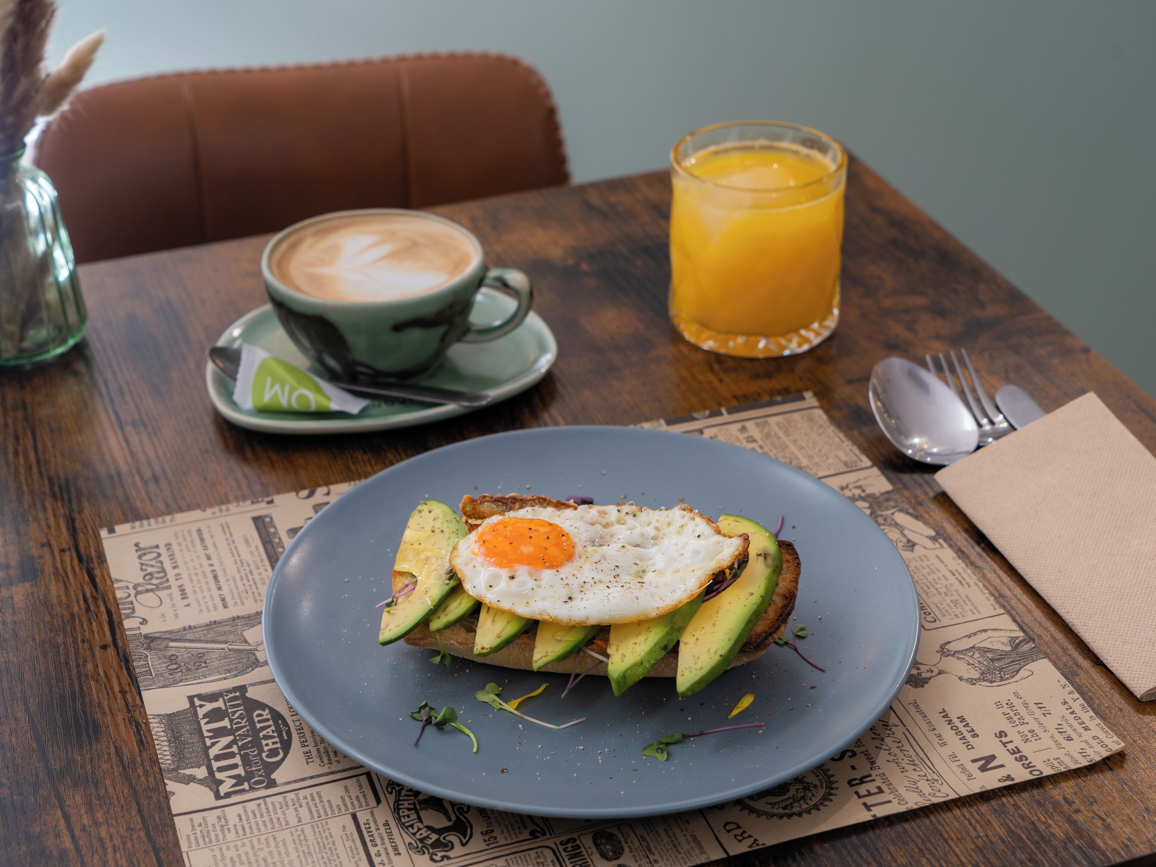 A breakfast plate with avocado toast and a fried egg, a cup of coffee, and a glass of orange juice on a wooden table.