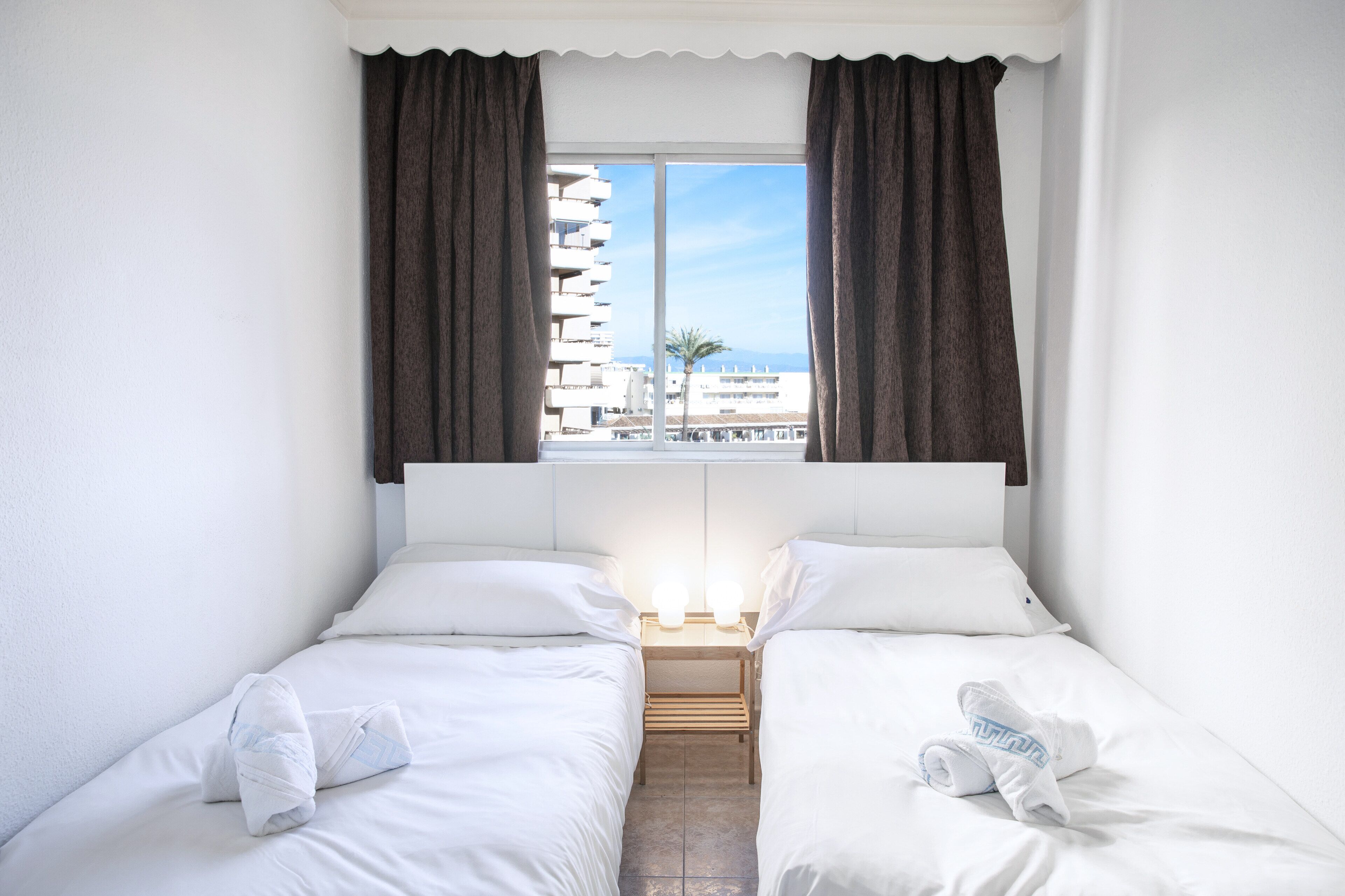 A hotel room with two single beds, a nightstand, a window with curtains, and a view of a building and palm tree.