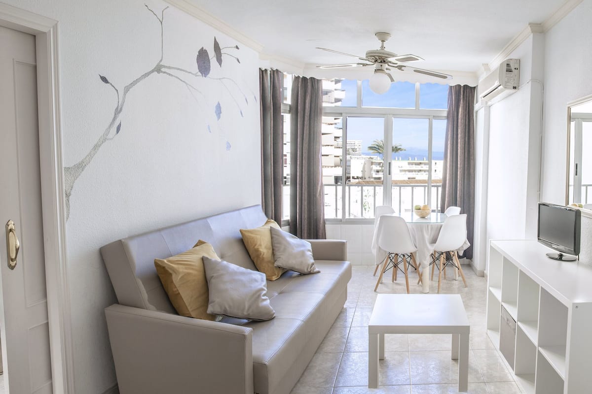A modern living room with a sofa, a coffee table, a TV, and a balcony with a view.