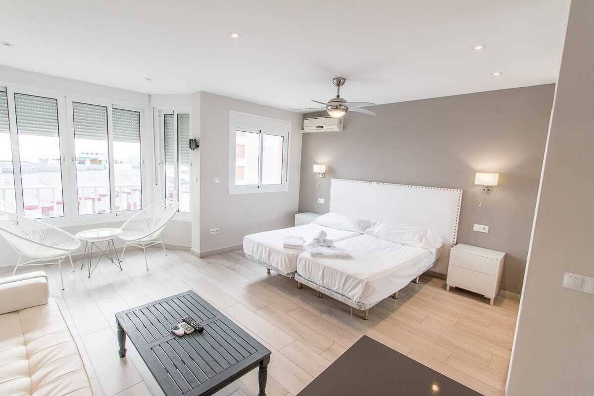 A modern hotel room with two beds, a balcony with white chairs, a small table, and a ceiling fan.