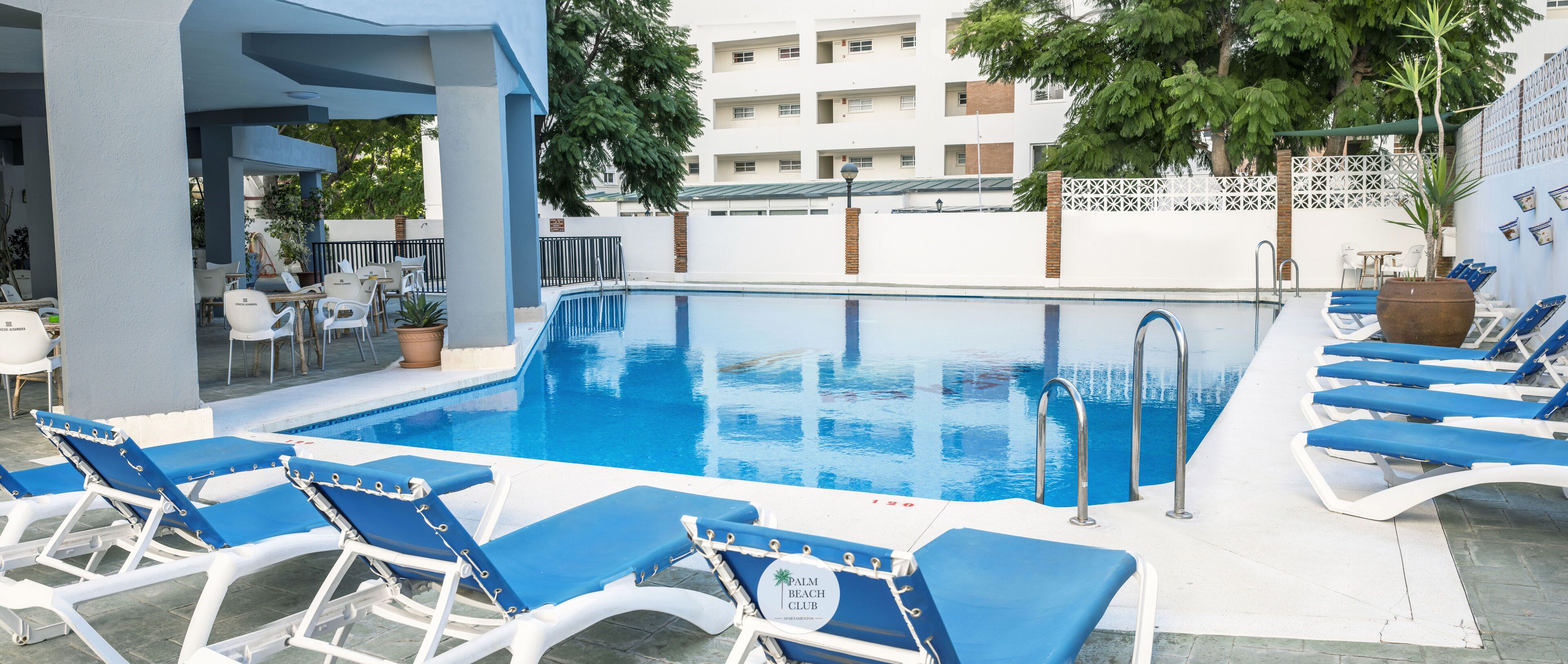 A pool area with blue lounge chairs, a pool, and a building in the background.