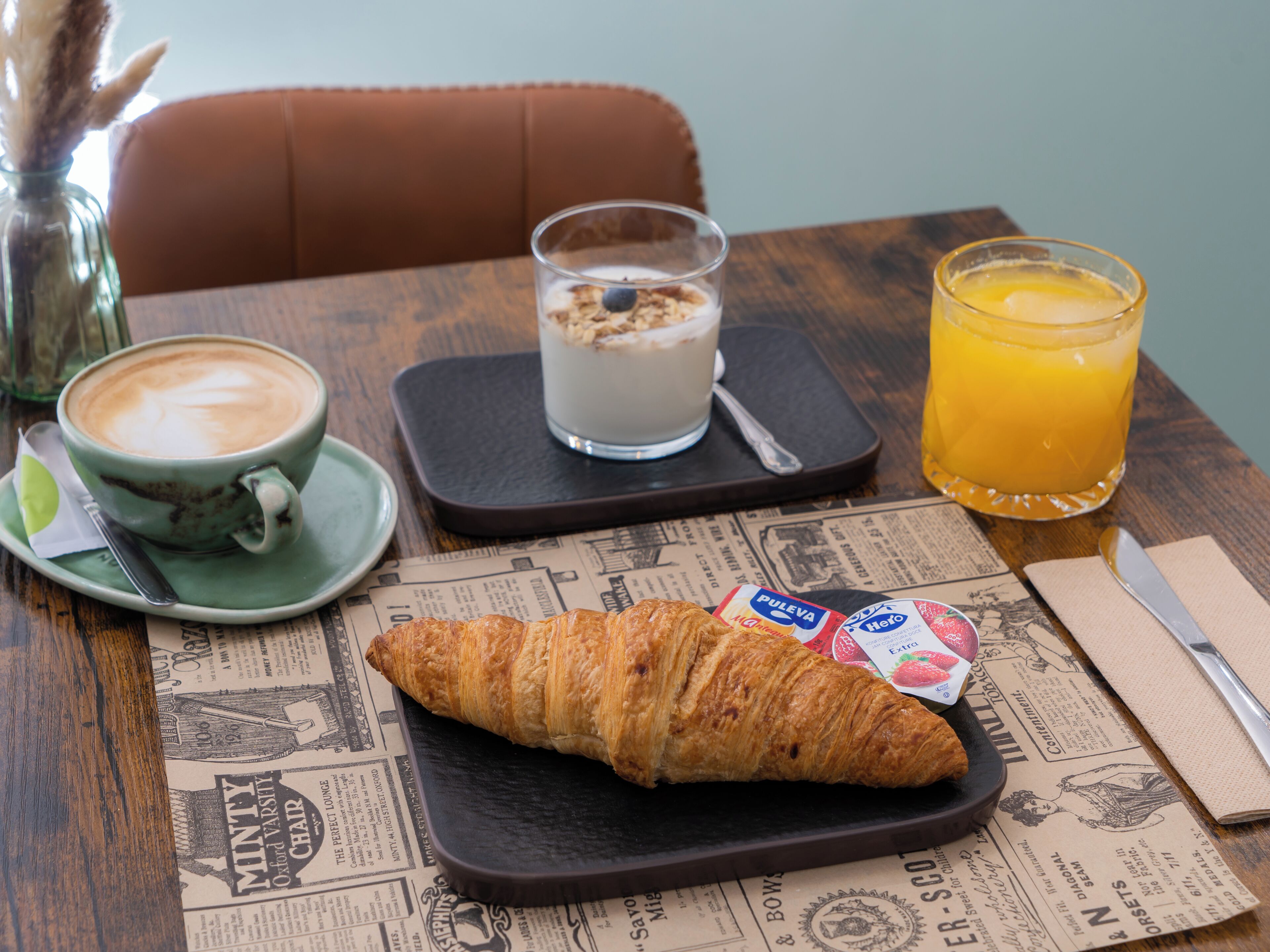 A breakfast setting with a croissant, a glass of orange juice, a cup of coffee, and a glass of milk with granola on a newspaper placemat.