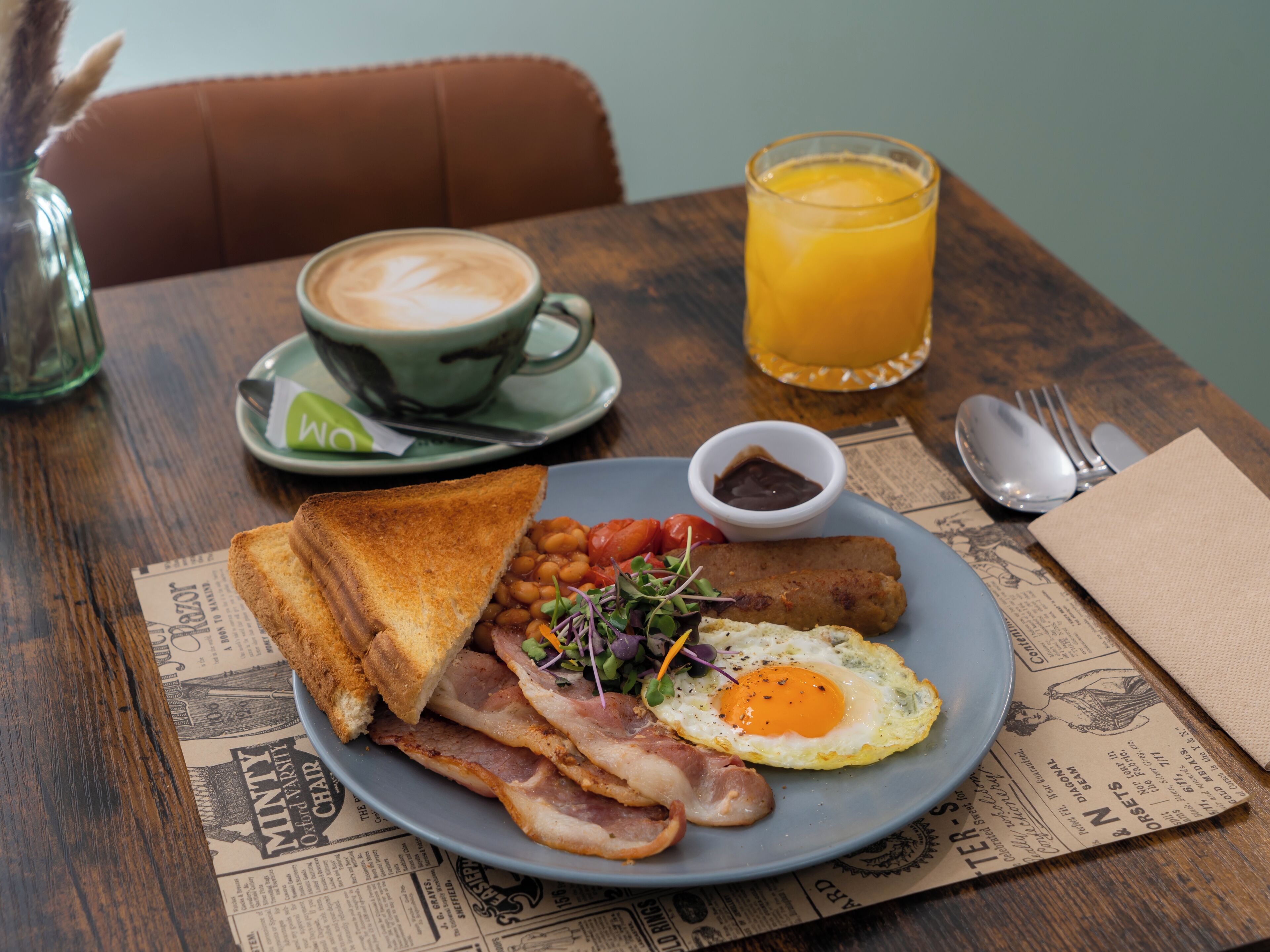 A breakfast plate with toast, bacon, eggs, beans, and a small salad, a cup of coffee, and a glass of orange juice on a wooden table.