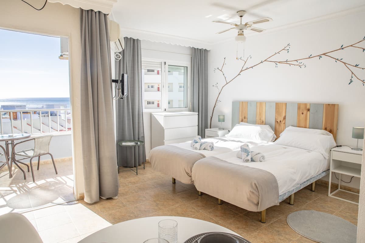 A hotel room with two beds, a balcony, a desk, and a view of the ocean.