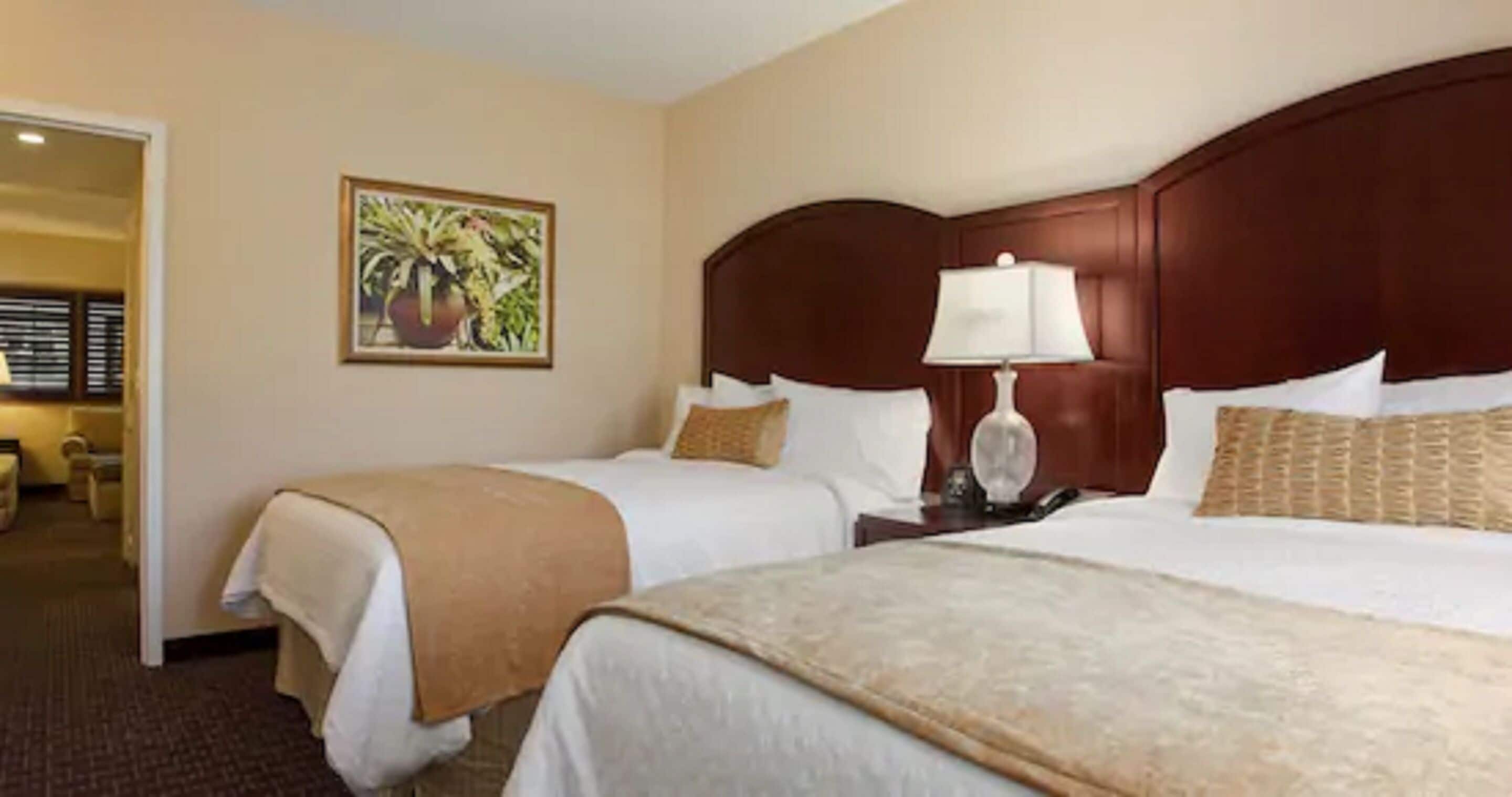 A hotel room with two beds, a wooden headboard, a bedside table with a lamp, and a painting on the wall.