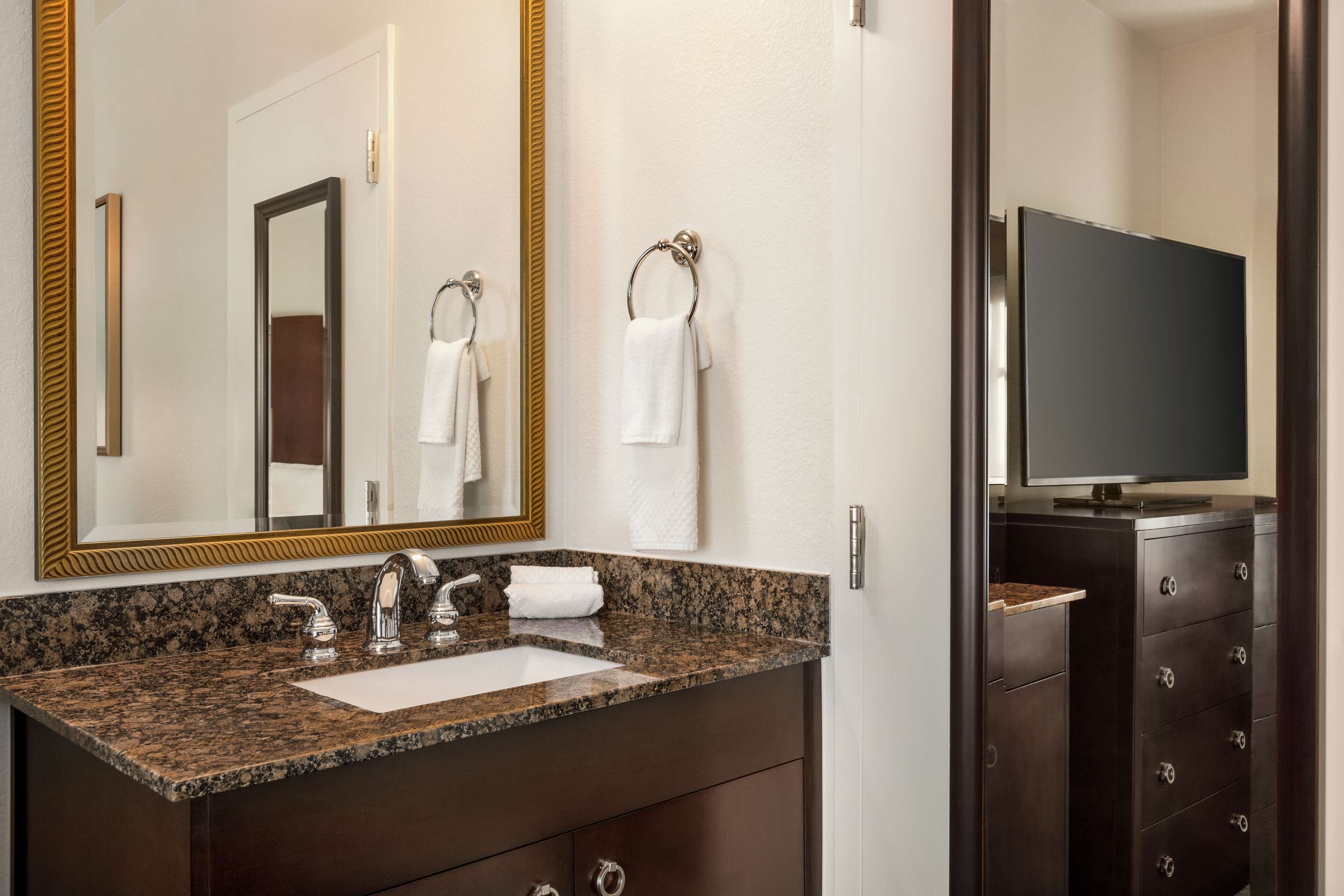A bathroom with a granite countertop sink, two faucets, a mirror with a gold frame, a towel rack, and a television on a dark wooden cabinet.