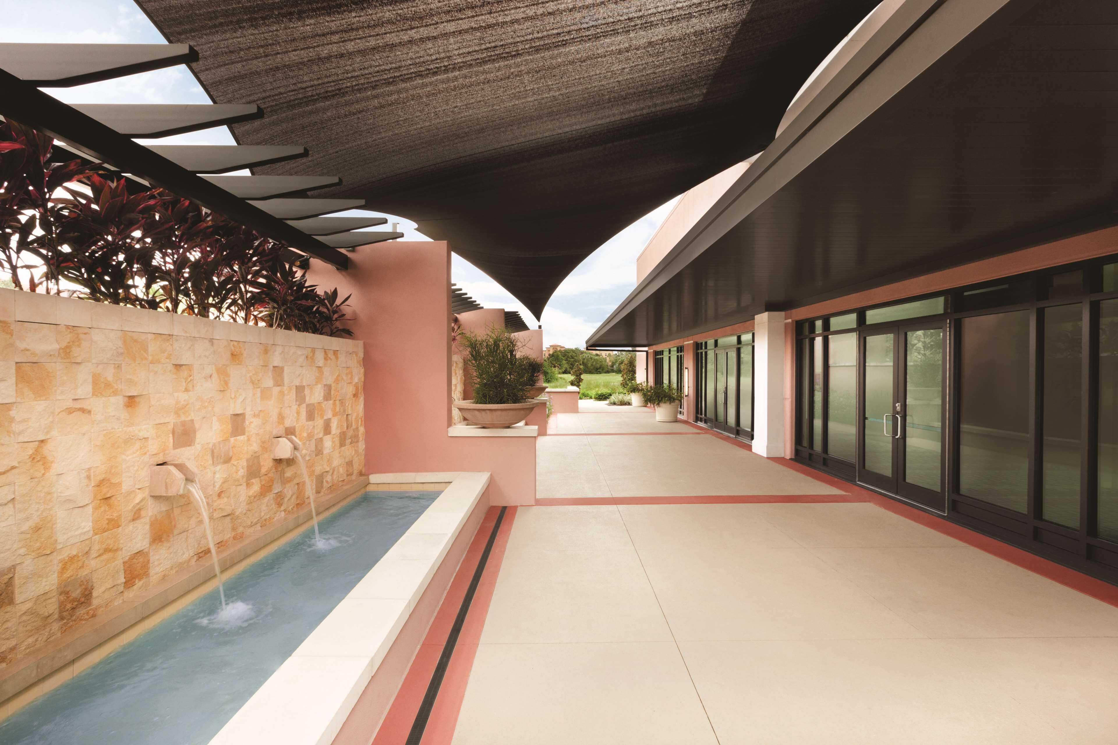 A modern outdoor pool area with a stone wall, two water features, and a covered walkway.