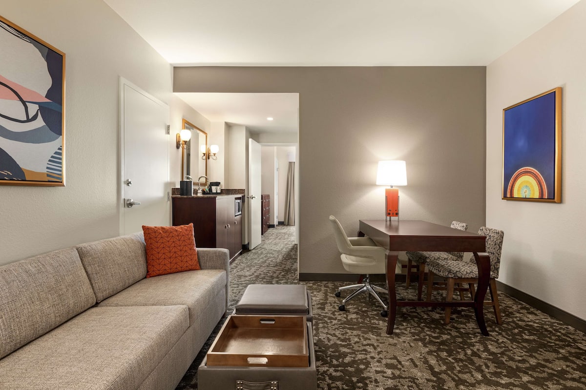 Habitación de hotel con sofá, una mesita, escritorio con silla, lámpara y obras de arte en las paredes.