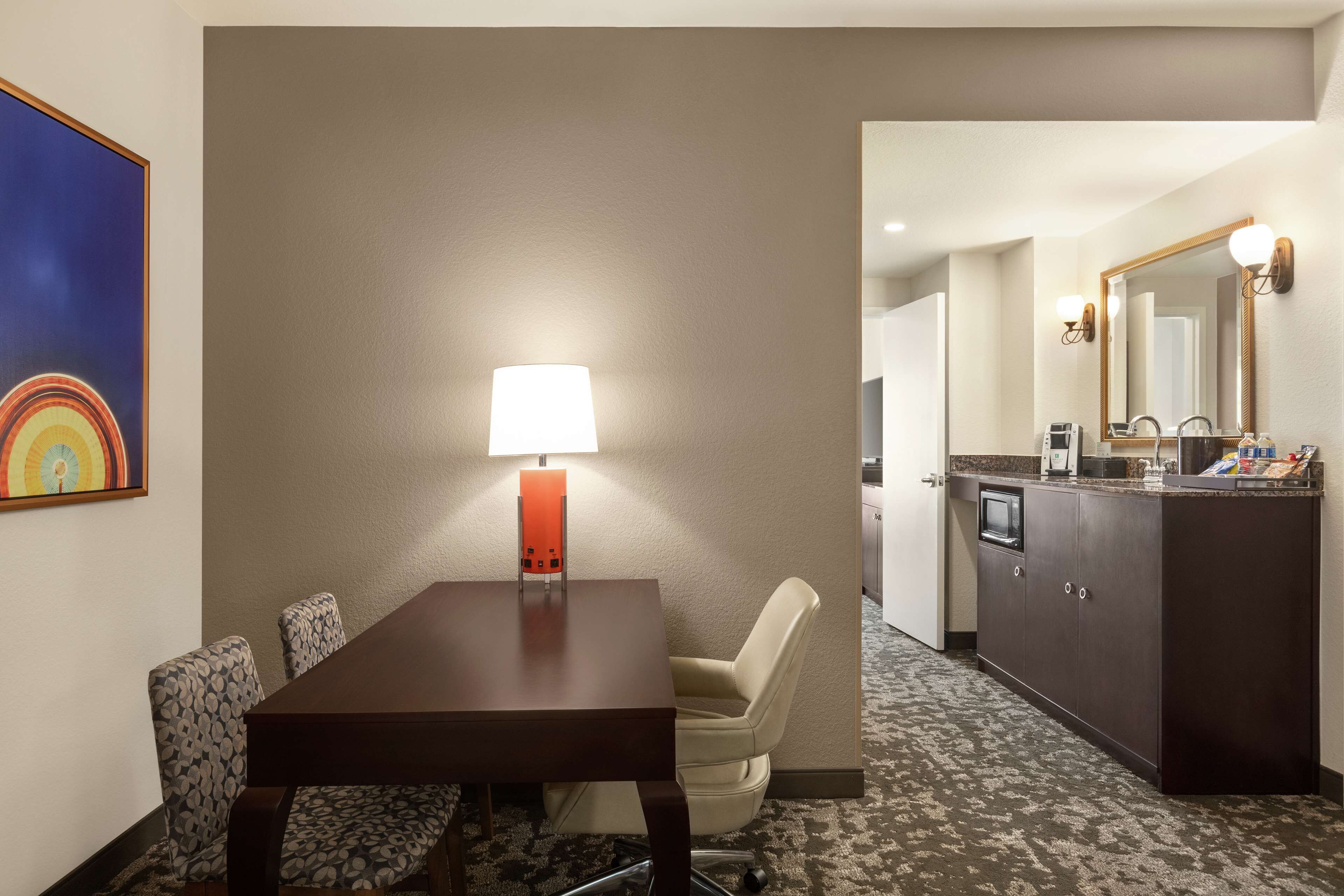 A hotel room with a wooden dining table, a single chair, a lamp, a kitchenette, and a large mirror.