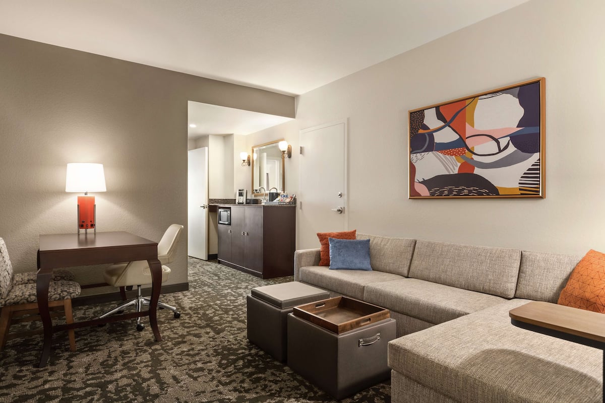 Habitación de hotel con sofá, escritorio con lámpara, kitchenette y un cuadro en la pared.