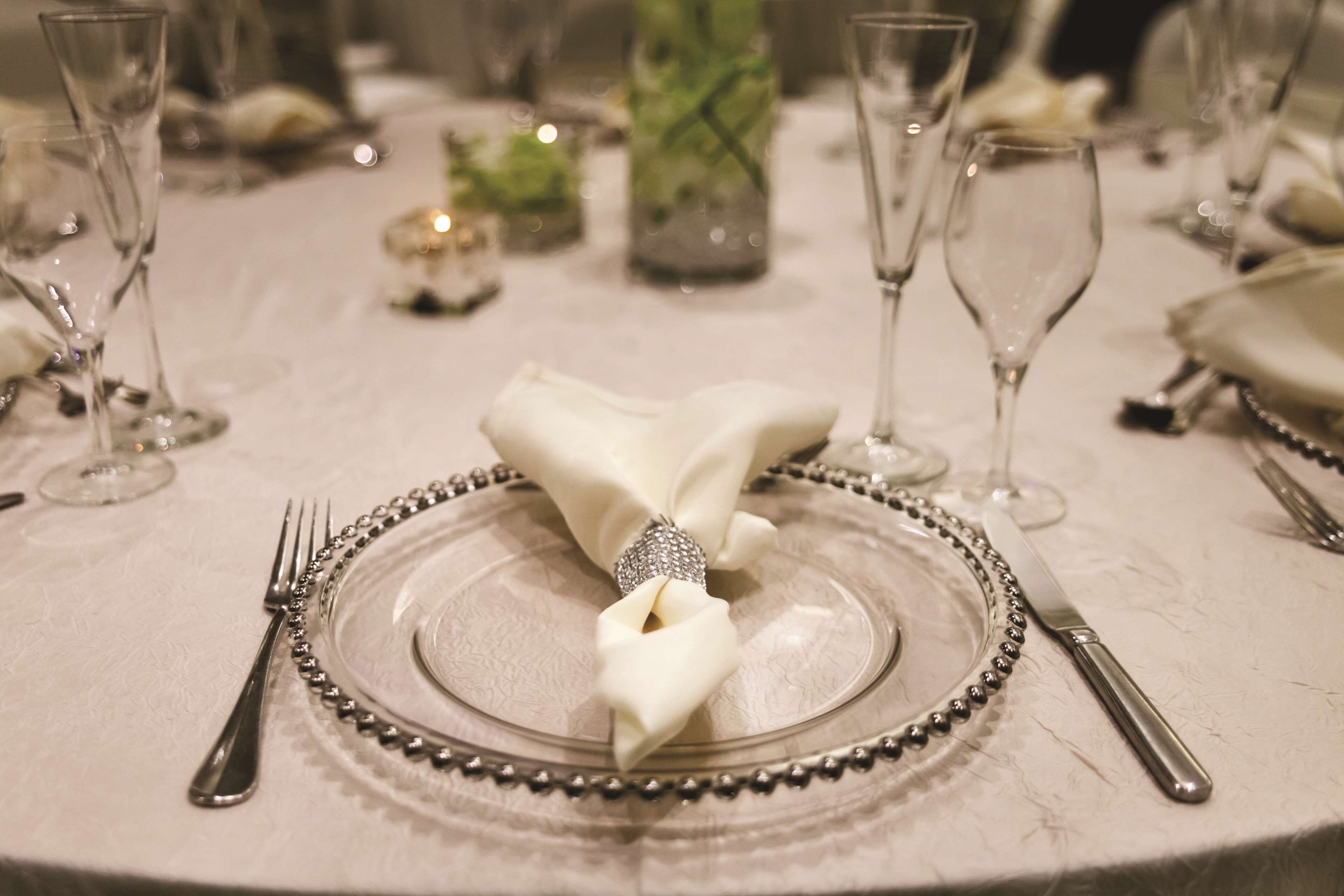 A table set for a formal dinner with a clear glass plate, silverware, and a folded napkin with a decorative band.