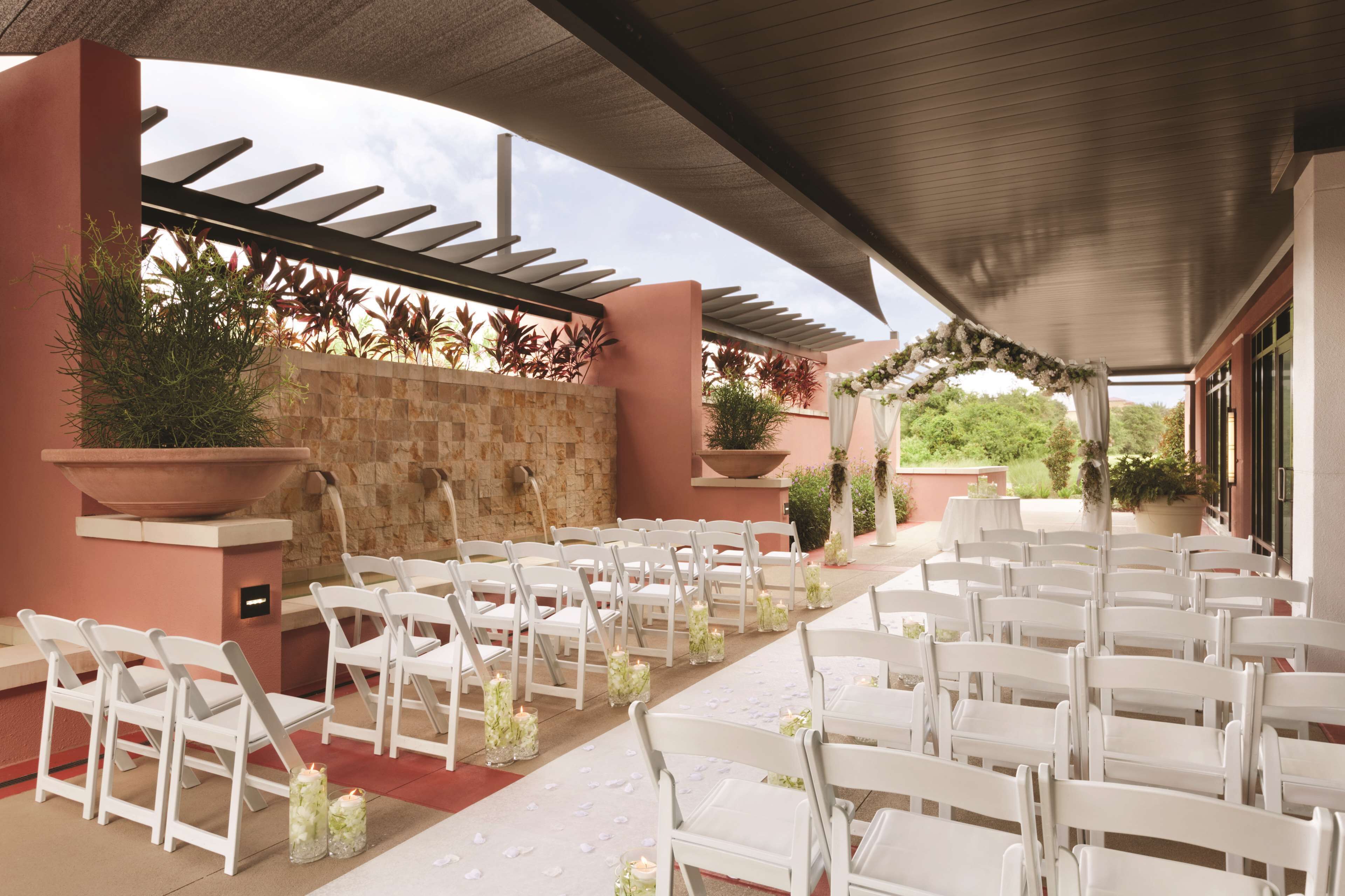 Un salón de bodas al aire libre con sillas y mesas blancas, paredes de piedra y plantas en macetas.