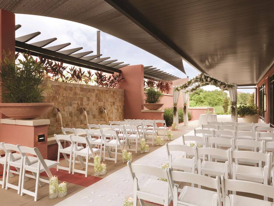 An outdoor wedding venue with white chairs and tables, stone walls, and potted plants.