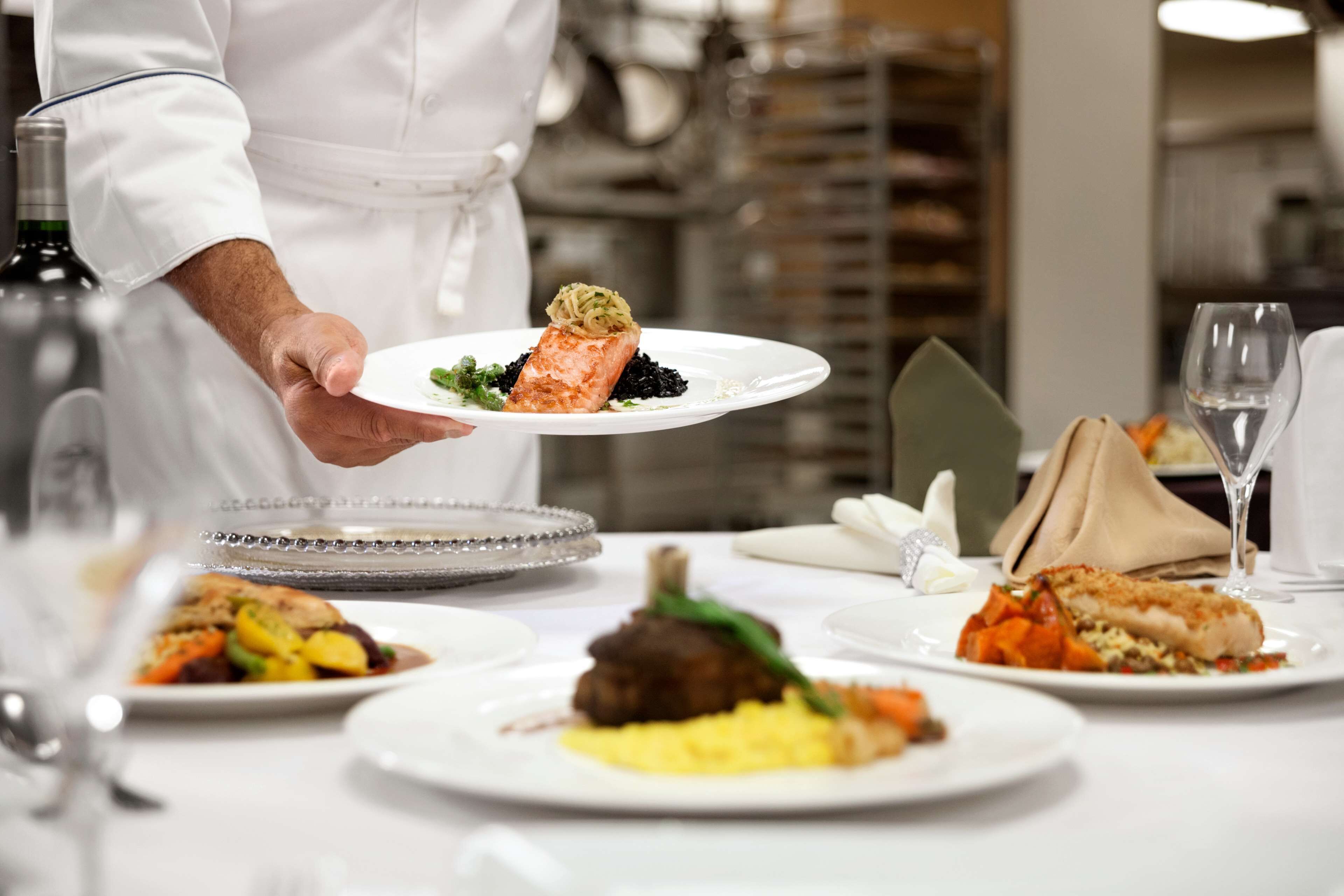 A chef serving a dish at a restaurant table with various other dishes and a wine glass.