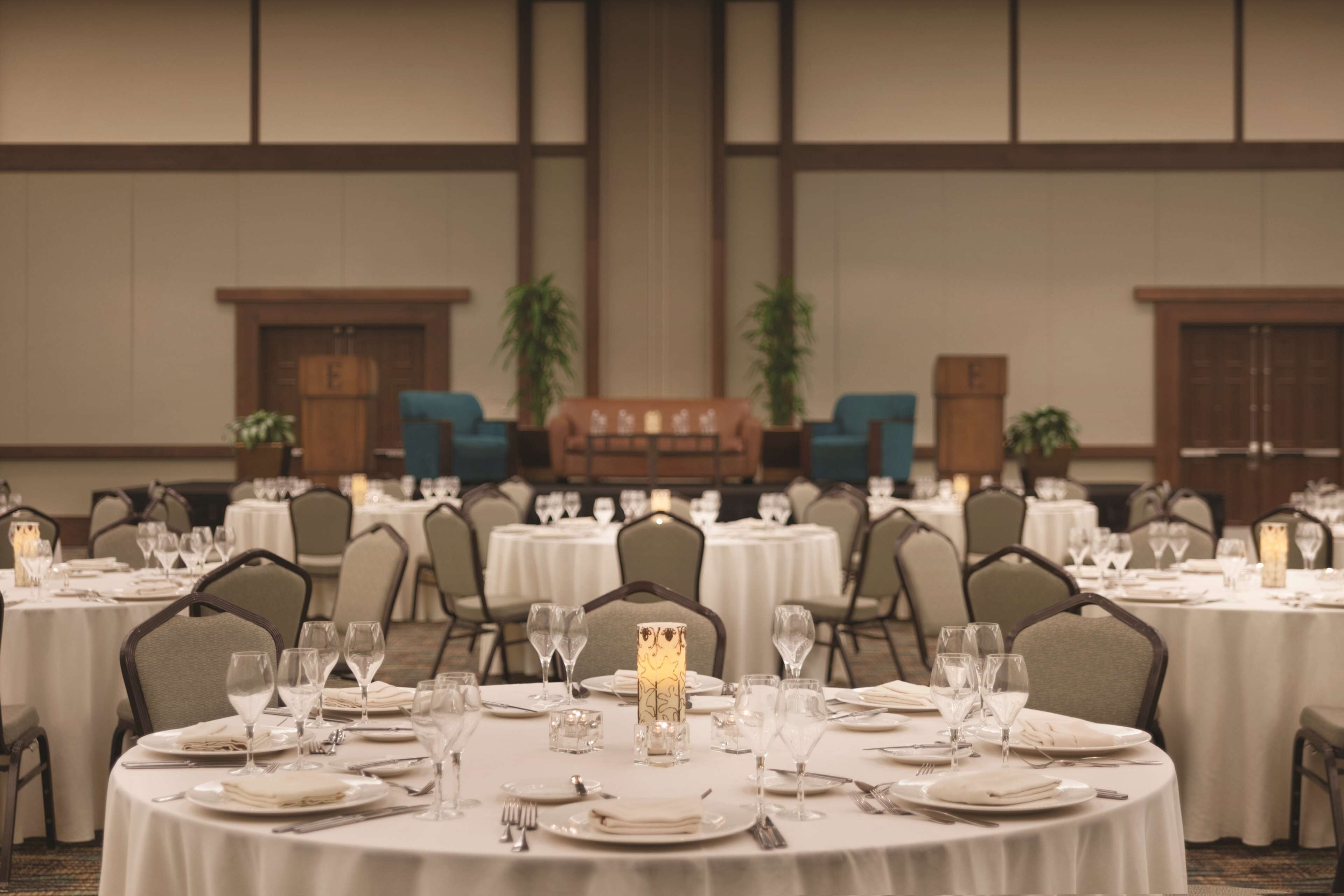 Un salón de banquetes con mesas redondas dispuestas para comer, cada una con mantel blanco, platos, cubiertos y copas.