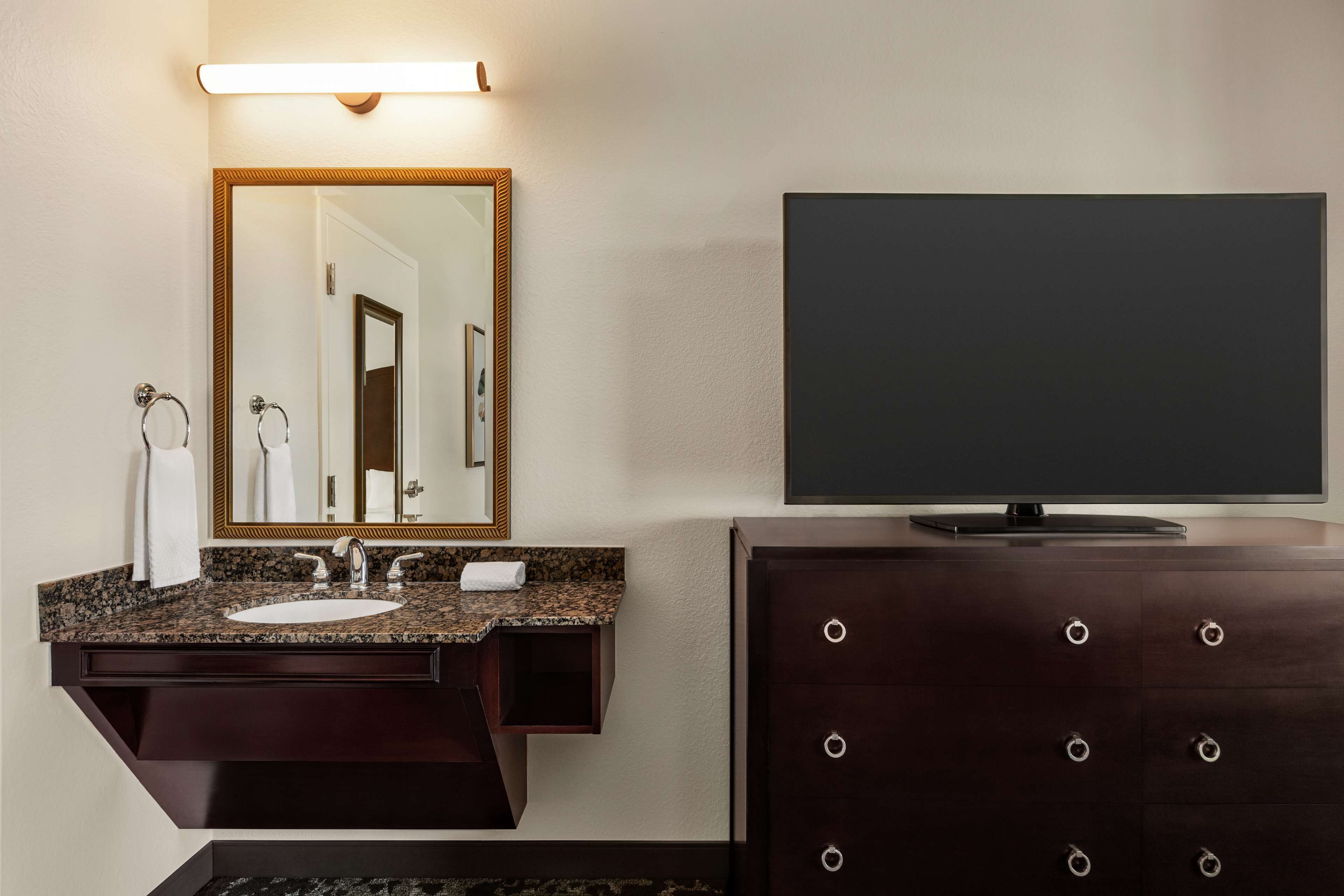 Un baño con lavamanos de granito, un espejo grande, un mueble de tocador de madera oscura con cajones y un televisor de pantalla plana montado sobre el mueble.