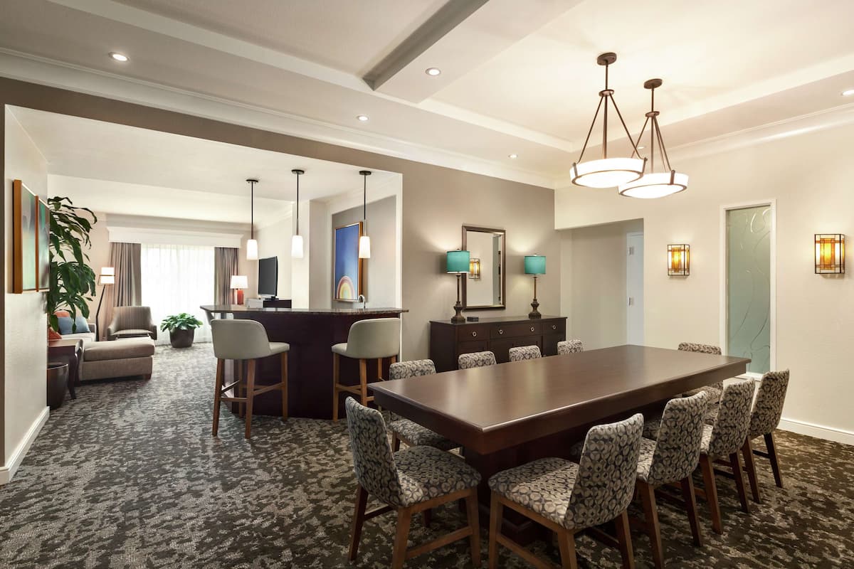Habitación de hotel con una amplia mesa de comedor, varias sillas, un sofá, un escritorio y una planta.