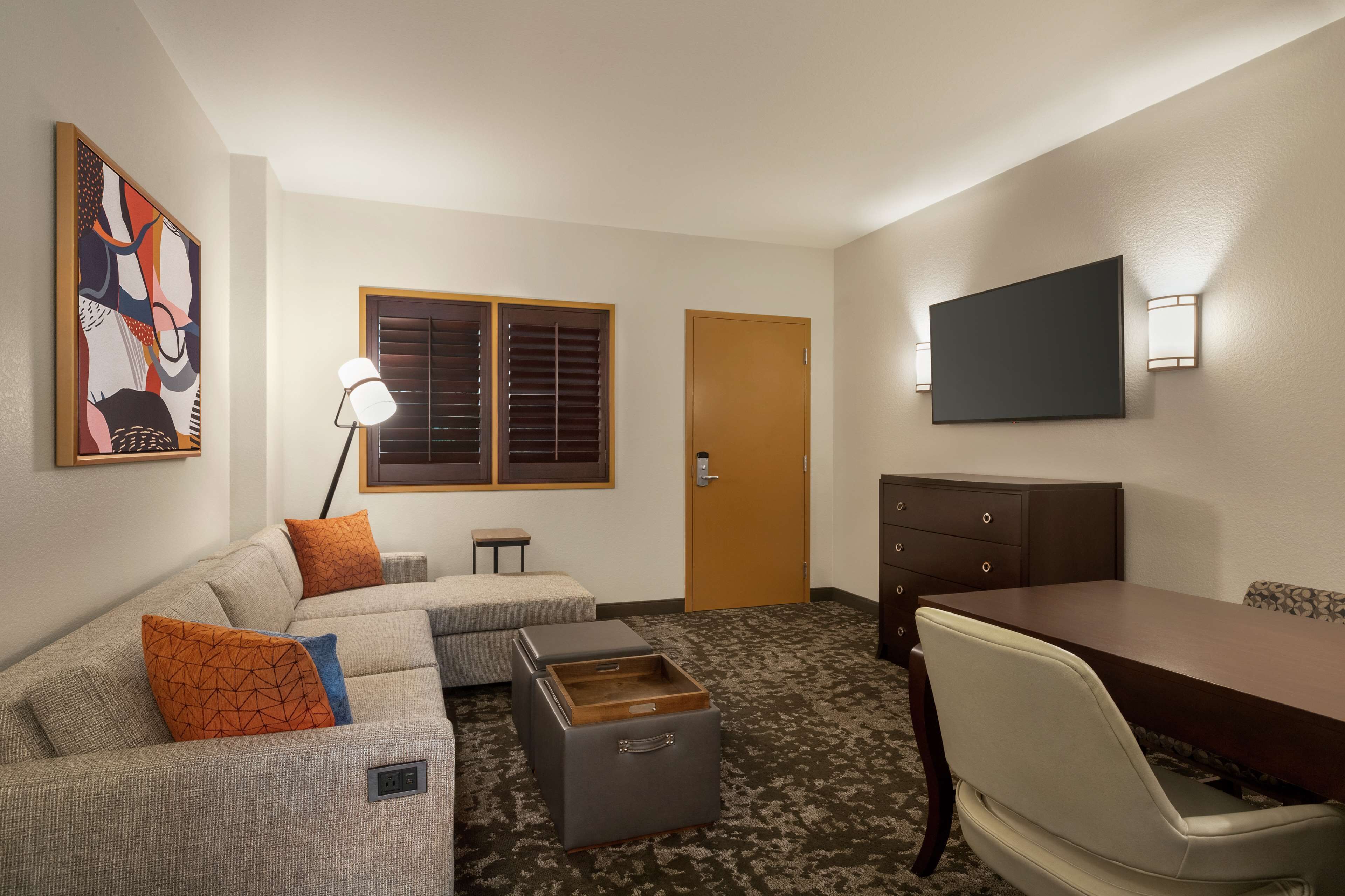 A hotel room with a sofa, ottoman, a small table, a desk, a chair, a TV, and a door.