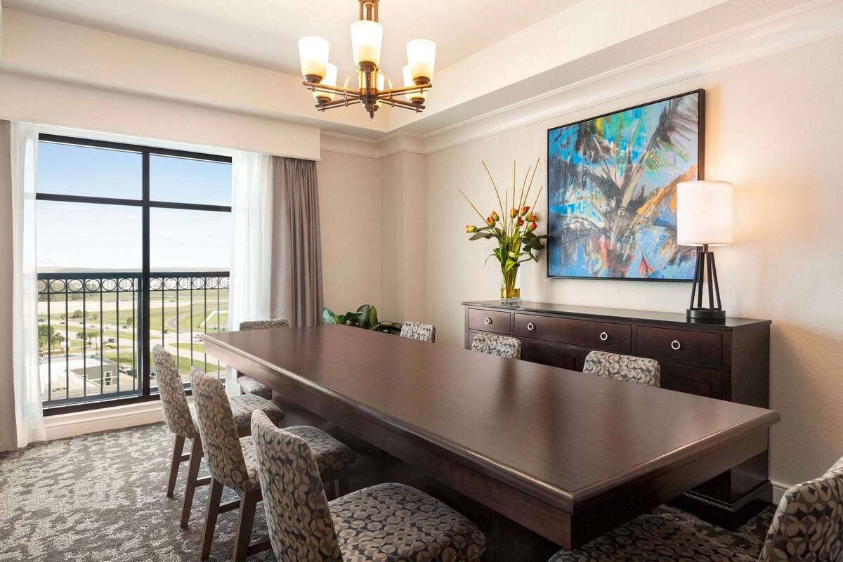 Un comedor de hotel con una mesa grande, seis sillas estampadas, un candelabro, un cuadro, un jarrón con flores y vistas a través de un ventanal.