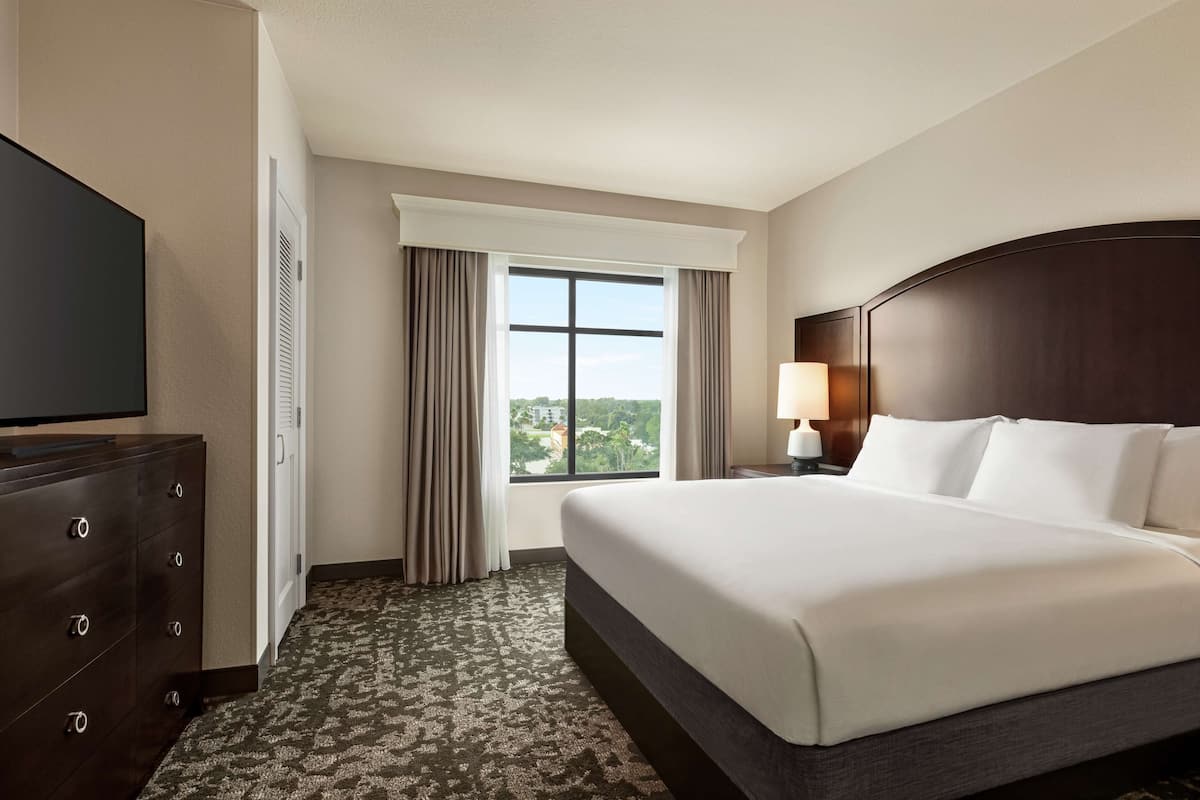 Habitación de hotel con una cama grande, un aparador, un televisor, una ventana con cortinas y una lámpara.