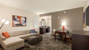 A hotel room with a sofa, ottoman, dining table, and chairs. There is a kitchenette in the background.