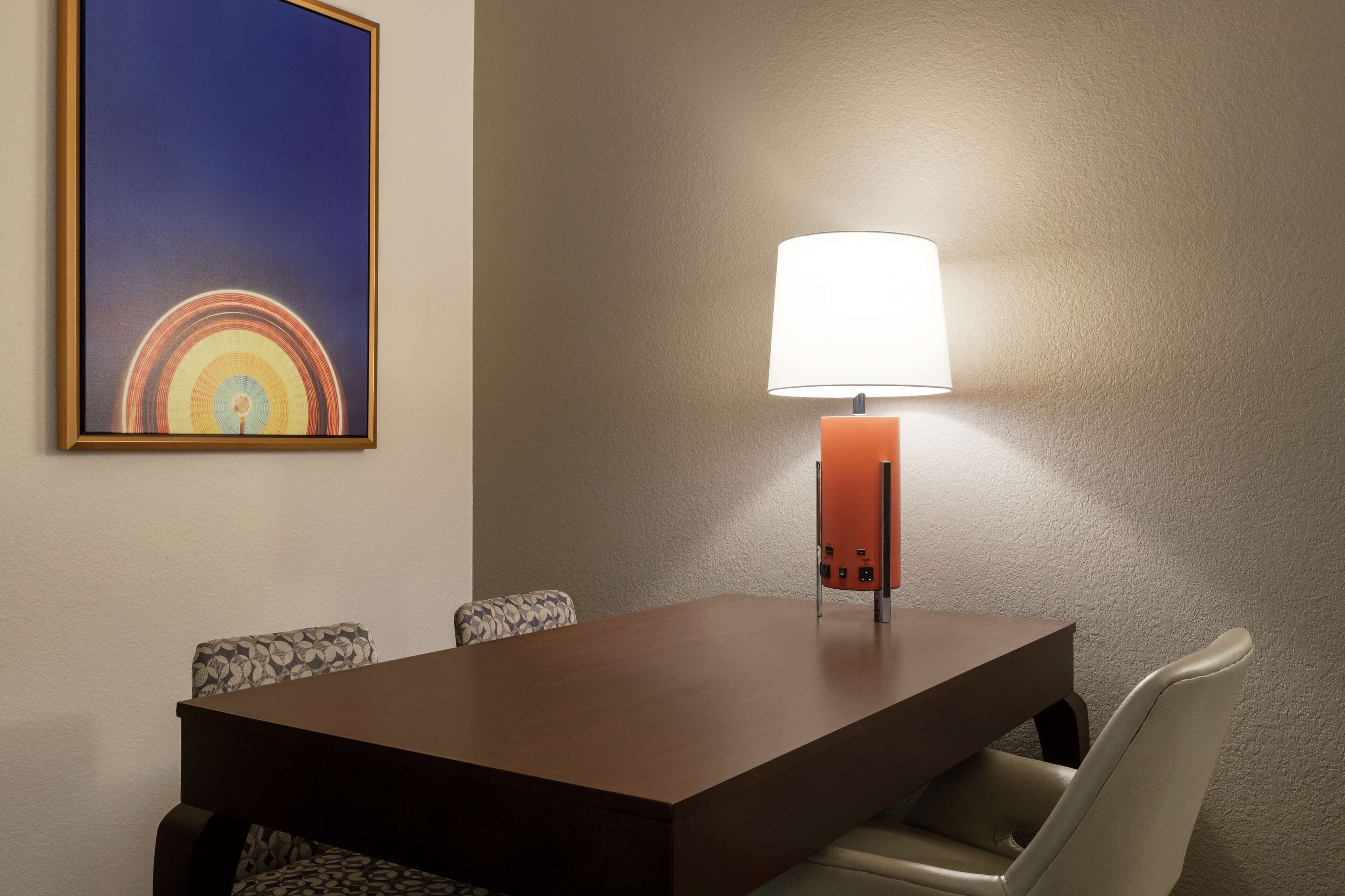 Een eetgedeelte met een tafel, stoelen, een lamp en een lijst met een afbeelding aan de muur.