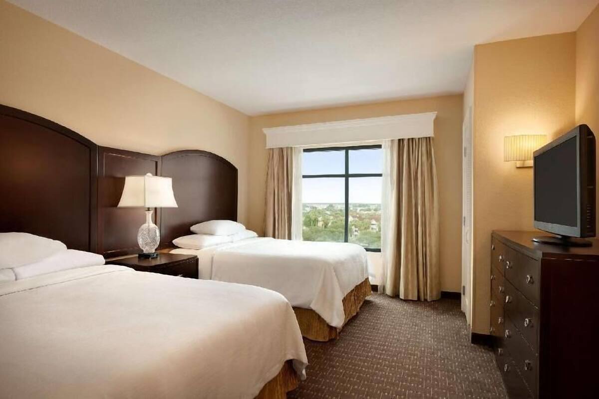 Ein Hotelzimmer mit zwei Betten, einem Fernseher, einer Lampe, einem Schminktisch und einem Fenster mit Vorhängen.