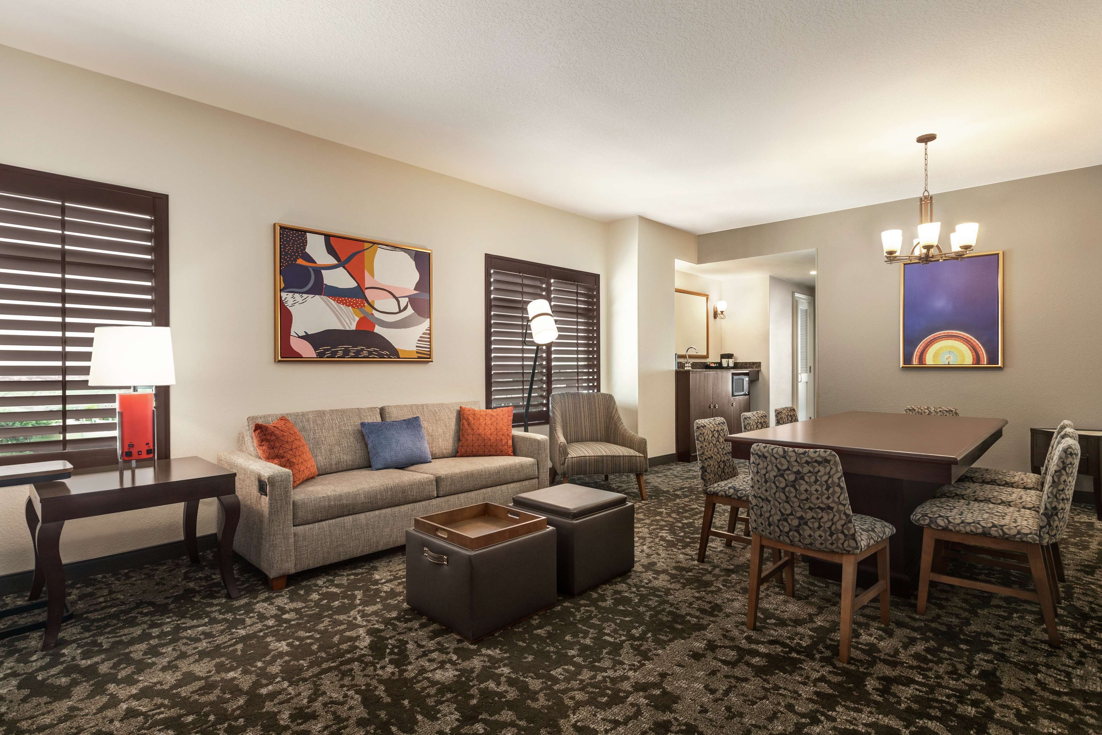 A hotel room with a sofa, ottomans, a dining table with chairs, a lamp, and artwork on the walls.