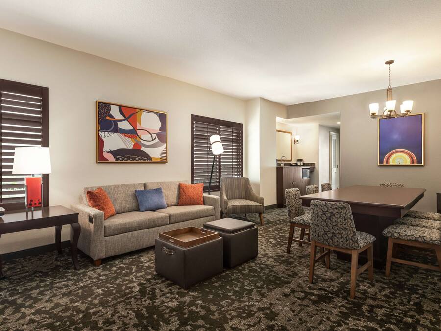 A hotel room with a sofa, ottomans, a dining table with chairs, a lamp, and artwork on the walls.