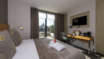 A hotel room with a large bed, a desk with a chair, a flat-screen TV, and a view of a cityscape through the window.