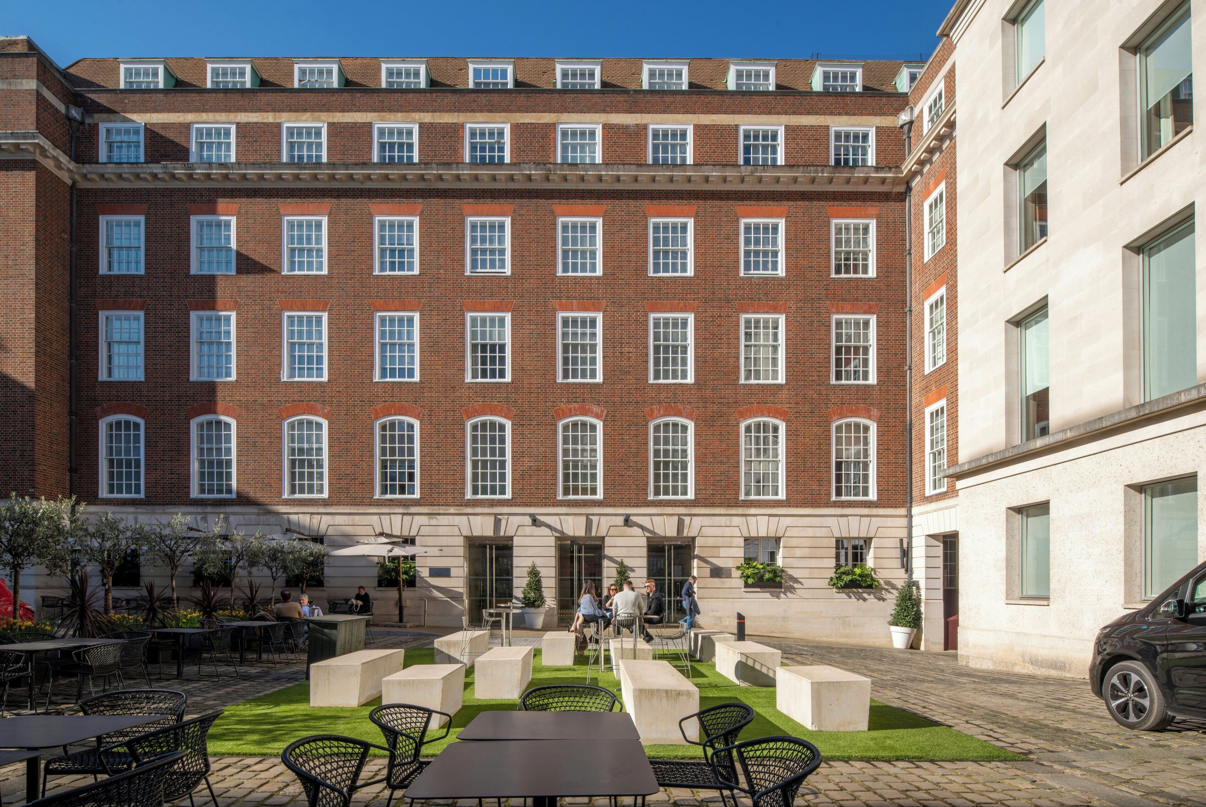 A courtyard with outdoor seating, a brick building with multiple windows, and a car parked nearby.
