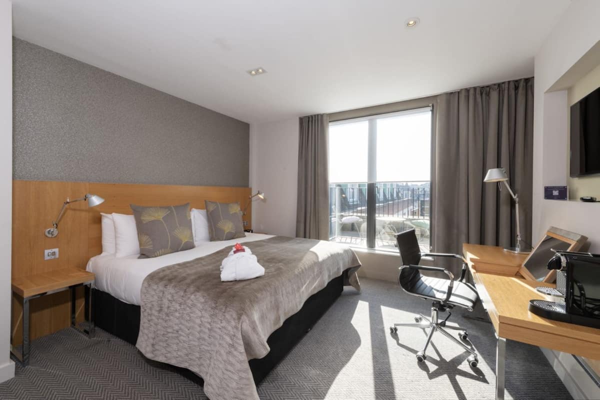 A hotel room with a large bed, a desk with a computer, a chair, and a view of the city through large windows.
