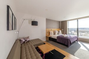 Ein modernes Hotelzimmer mit einer Couch, einem Bett mit lila Decke, einem Schreibtisch und einem großen Fenster mit Blick auf die Stadt.