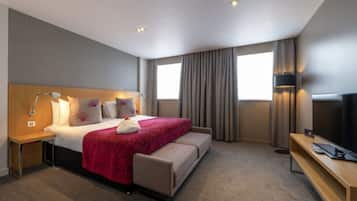 Quarto de hotel com cama, mesa de cabeceira, luminária, televisão e janela com cortinas.