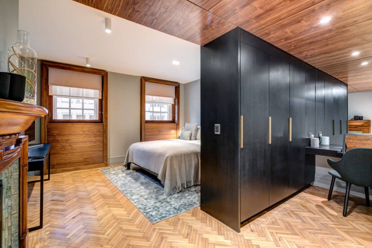 A modern bedroom with a wooden floor, a bed with a beige bedspread, a black wardrobe, and a small desk area.