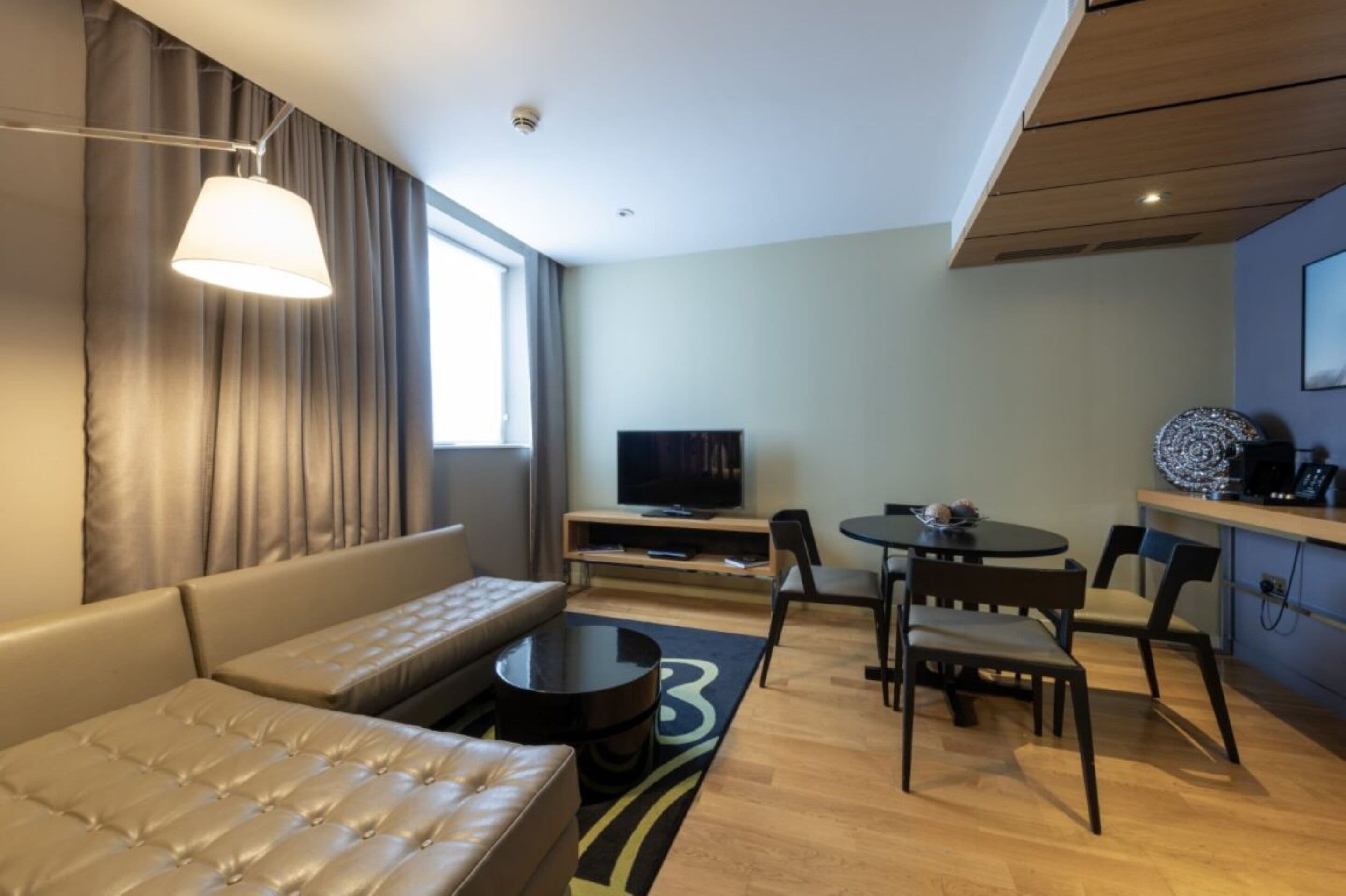 A modern hotel room with a sofa, a coffee table, a TV, and a dining area with chairs and a small table.
