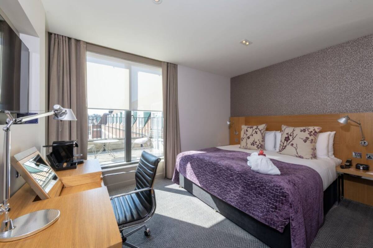 A hotel room with a bed, desk, chair, and a view of the outside.