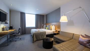 Ein Hotelzimmer mit Bett, Schreibtisch mit Laptop, Sofa, Stehlampe und einem großen Fenster mit Vorhängen.