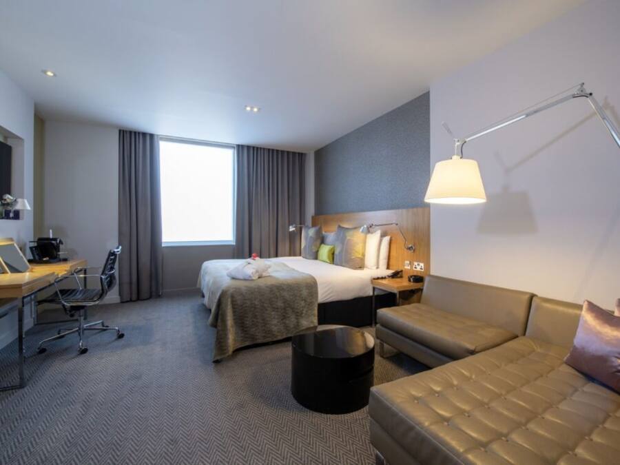 A hotel room with a bed, a desk with a laptop, a sofa, a standing lamp, and a large window with curtains.