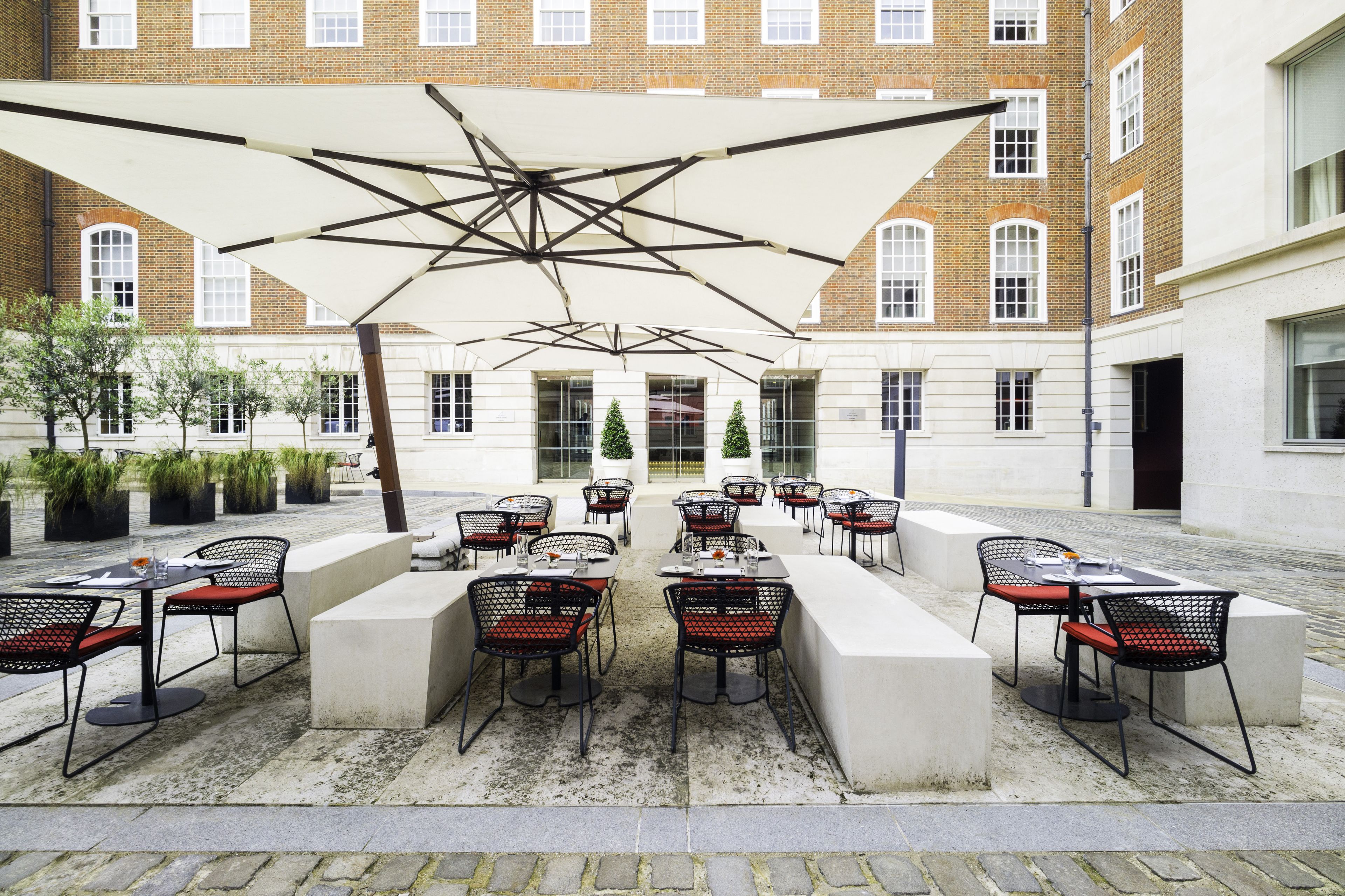 An outdoor cafe with tables and chairs, a large umbrella, and a building with brick walls in the background.