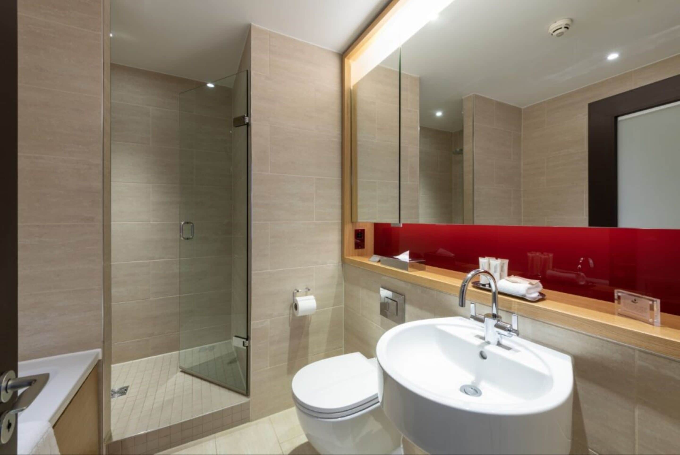 Une salle de bain moderne avec une douche à parois de verre, un lavabo blanc et un mur d’accent rouge.