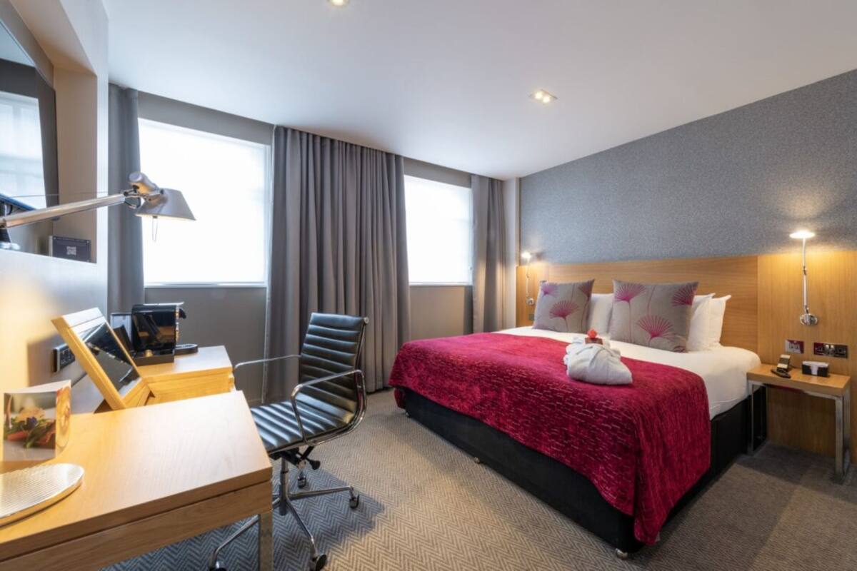 A hotel room with a bed, a desk with a computer, a chair, a desk lamp, and a nightstand with a lamp.