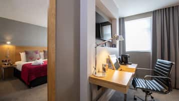 Habitación de hotel con cama, escritorio, silla, lámpara y un ventanal con cortinas.