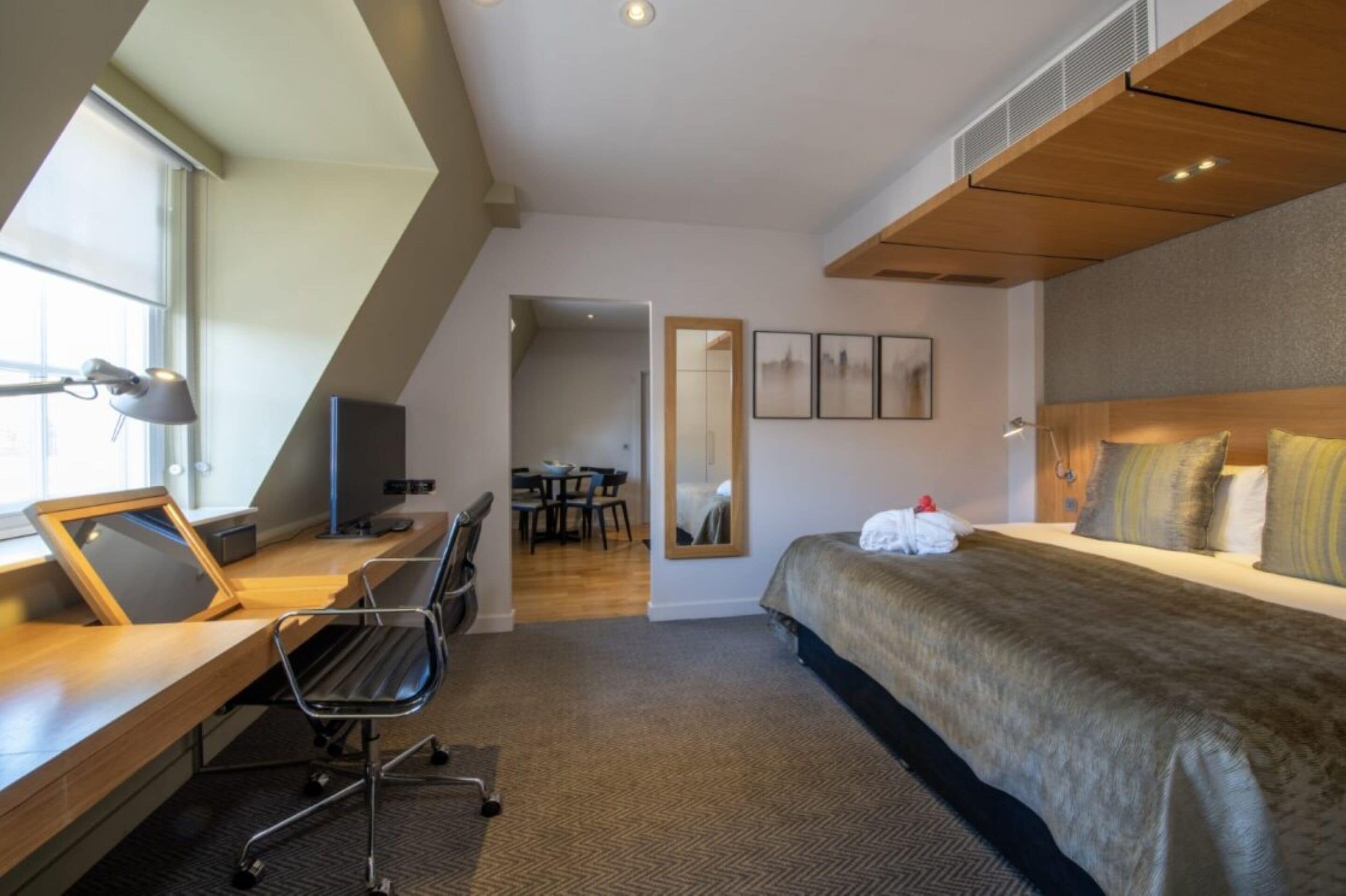 Ett modernt hotellrum med en stor säng, ett skrivbord med en dator, en stol och en matplats som syns genom en öppen dörr.