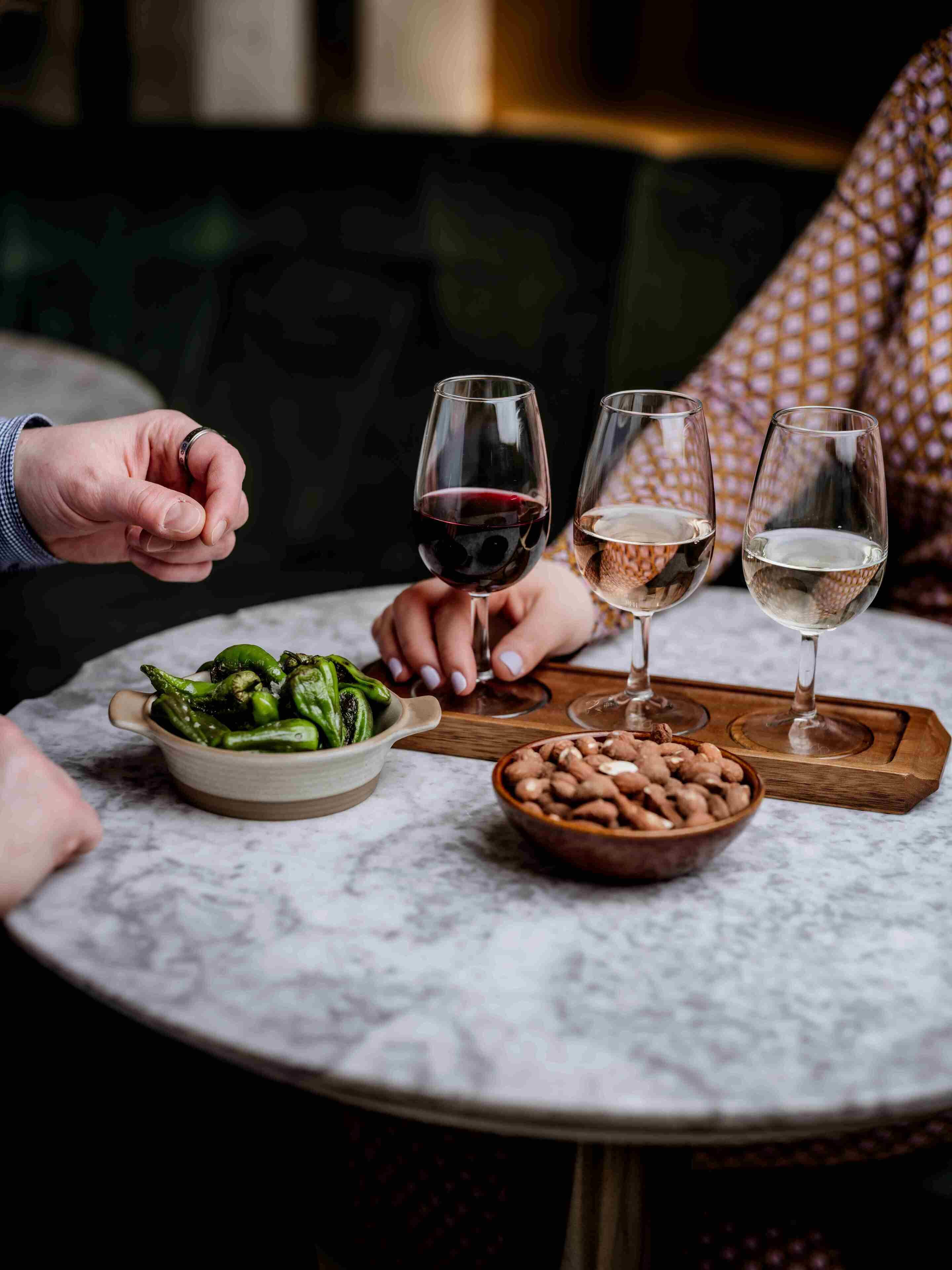 A round table with a marble top, two wine glasses, a bowl of green vegetables, and a bowl of nuts.